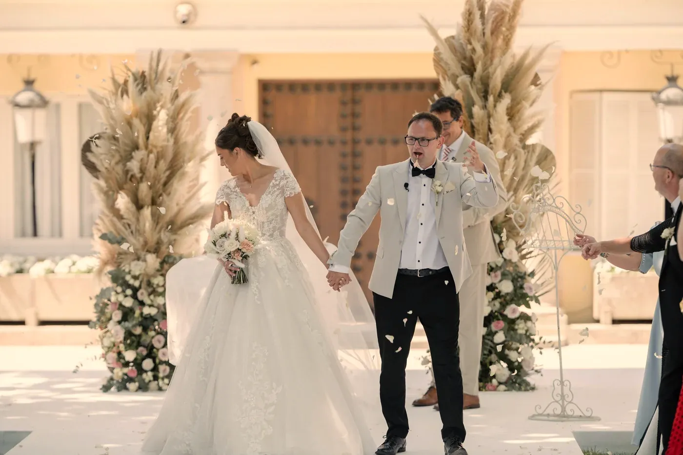 A couple walking back down the aisle at an outdoor ceremony in front of a sunlit villa, with pampas grass arrangements