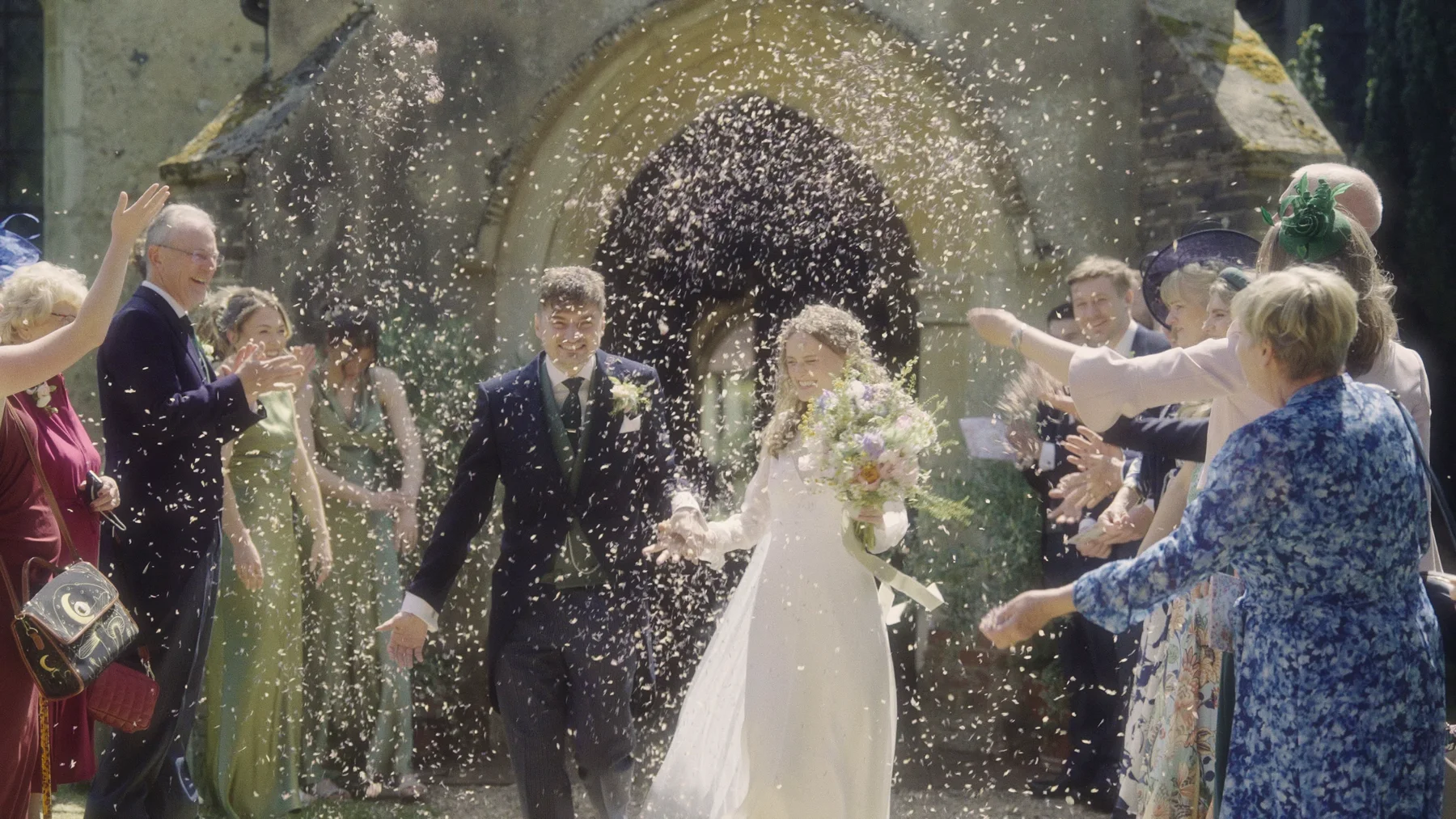 Charlotte and Dom walking through confetti outside All Saints Church in Ashdon